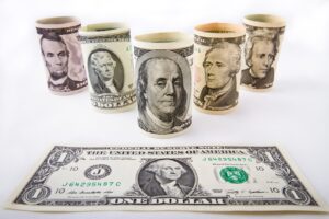 Cinco billetes de dólar estadounidense enrollados en forma de cilindros sobre un fondo blanco, con un billete de un dólar extendido en primer plano.