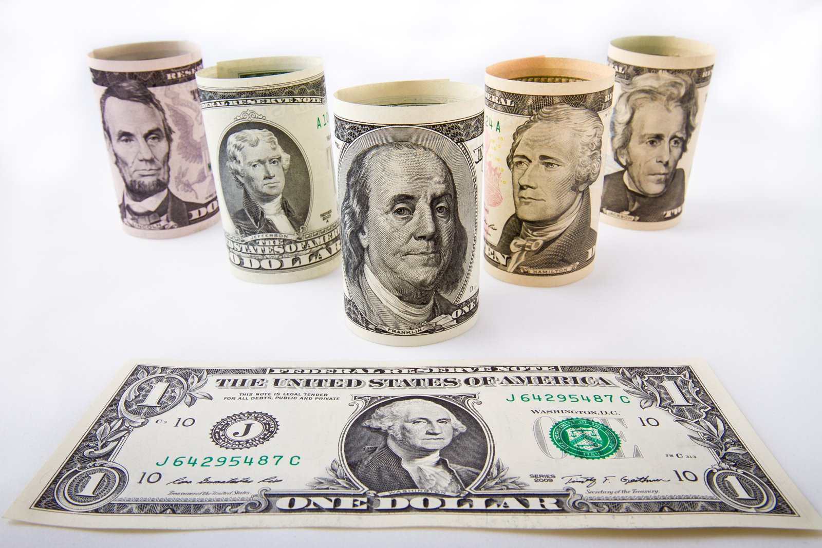 Cinco billetes de dólar estadounidense enrollados en forma de cilindros sobre un fondo blanco, con un billete de un dólar extendido en primer plano.