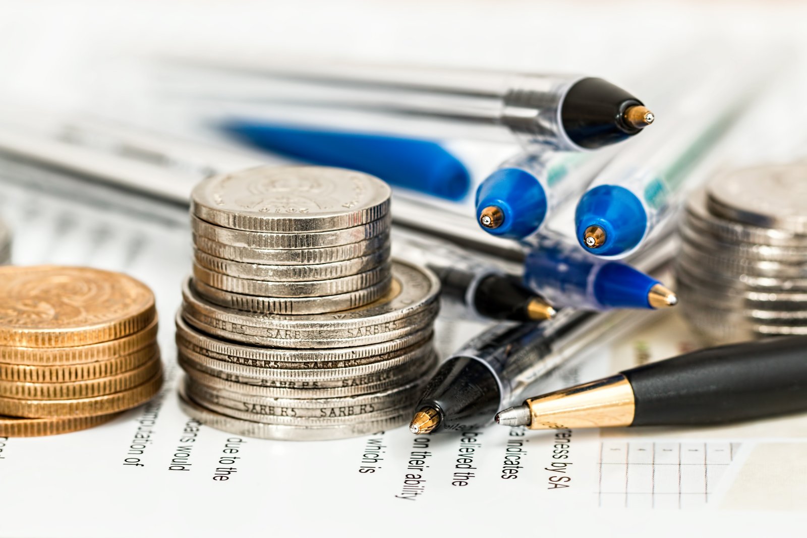 Pilas de monedas de plata y bronce junto a bolígrafos sobre documentos impresos con texto y una tabla, en una mesa.