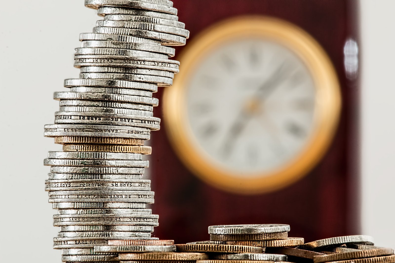 Pila vertical de monedas de plata junto a un reloj desenfocado al fondo, sobre una superficie clara.