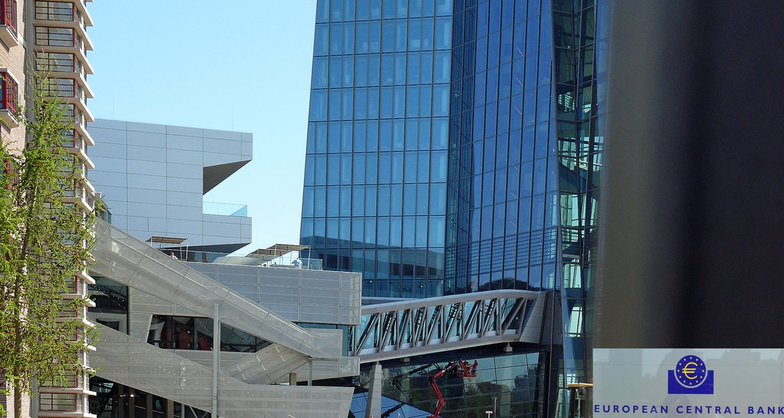 Edificio de oficinas de cristal azul con una pasarela elevada y una estructura moderna, junto a un logotipo del Banco Central Europeo en la esquina inferior derecha.