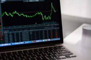 Primer plano de una laptop con una plataforma de trading abierta mostrando un gráfico de velas y una tabla de operaciones con comisiones y ganancias.