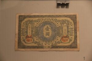 Un billete antiguo chino enmarcado y colgado en una pared, con caracteres y sellos rojos, sujeto por un clip metálico.