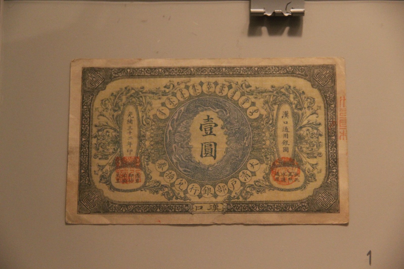 Un billete antiguo chino enmarcado y colgado en una pared, con caracteres y sellos rojos, sujeto por un clip metálico.